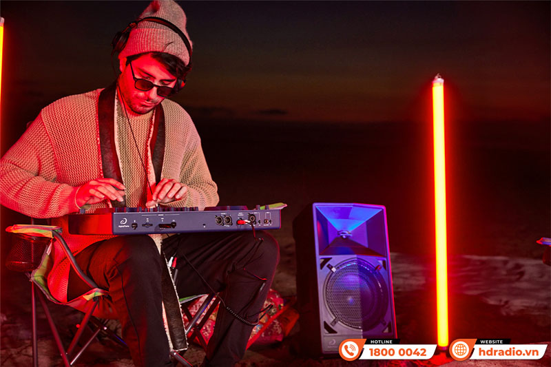 Buổi trình diễn âm nhạc ngoài trời vào ban đêm với DJ mặc áo len và đội mũ len, đang chơi bộ điều khiển DJ OMNIS-DUO cùng hệ thống loa và đèn LED sáng chói.