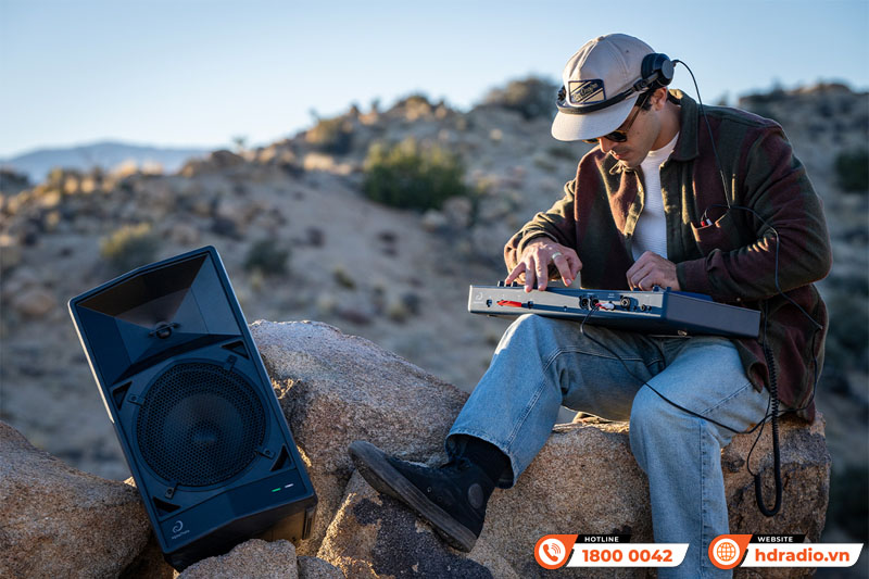 Người đam mê âm nhạc đang chơi nhạc điện tử trên bàn DJ di động giữa khung cảnh đồi núi, cạnh loa lớn đặt trên đá.